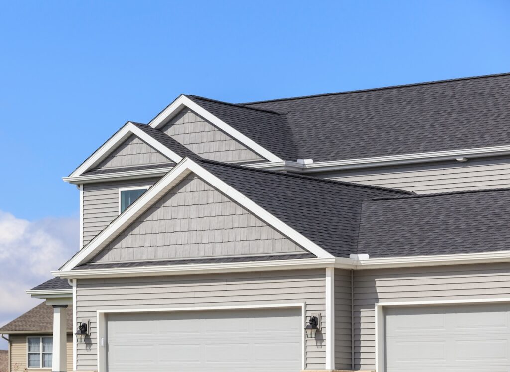 Close-up view of the upper portion of a modern suburban house with light gray siding, white trim, and multiple gables—showcasing popular roof types like dark asphalt shingles—under a clear blue sky.