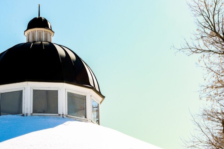 A black dome roof caps the white-walled cupola, both dusted with snow beneath a clear blue sky. Leafless tree branches frame the right side of the image.