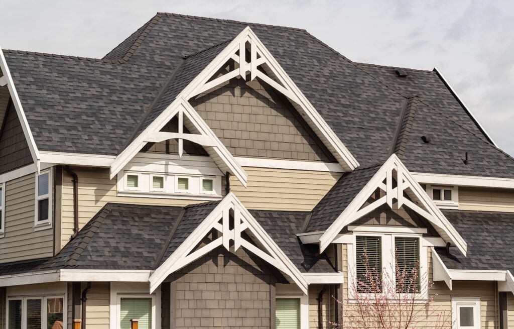 A close-up view of a house exterior showcasing multiple peaked gables, a roof type with dark gray shingles, beige and gray siding, and white trim accenting the windows and rooflines.