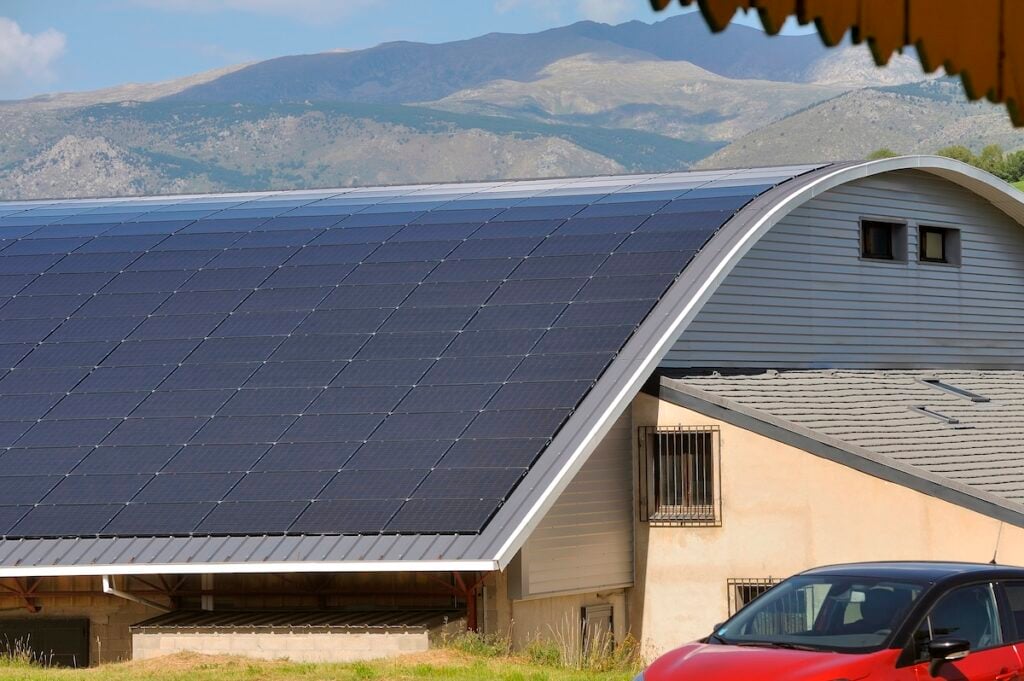 A large building with a striking curved roof covered in solar panels sits before mountains. The light-colored structure is complemented by a nearby red car, all beneath a clear, sunny sky.