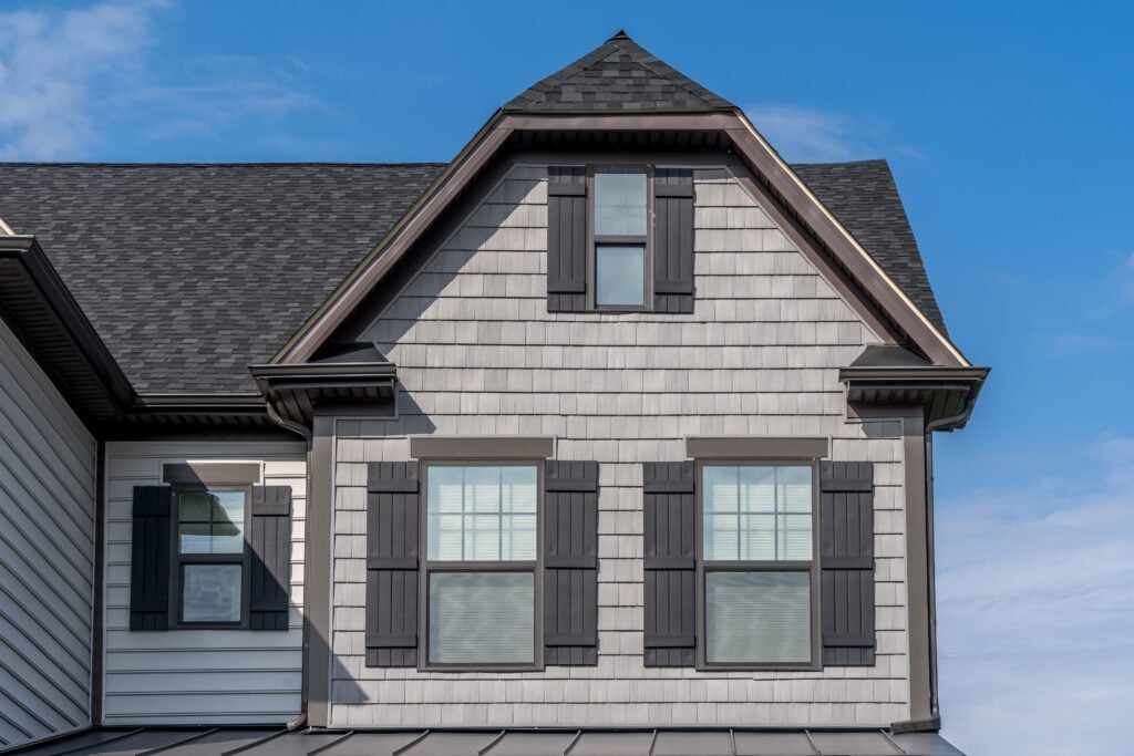 Close-up of the upper facade of a house with gray siding, black shutters, and three windows beneath a dark gray, steeply pitched dutch gable roof against a blue sky.