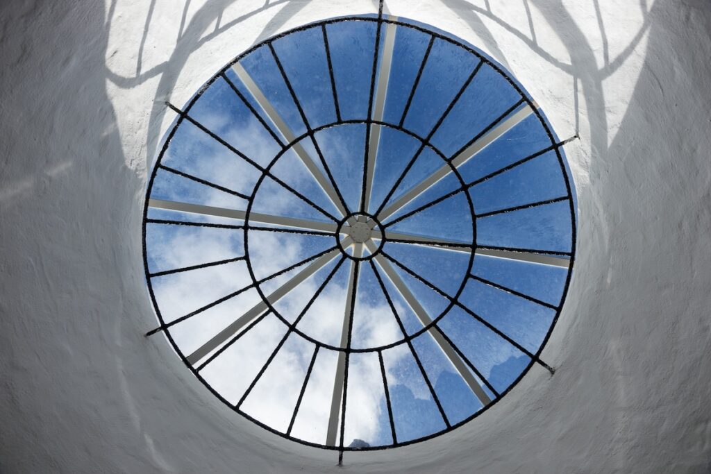 A round glass skylight with black metal spokes is set in a white ceiling, resembling a dome roof and showing a view of blue sky and clouds above. Sunlight and shadows create patterns around the opening.
