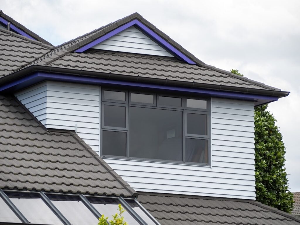 A close-up of the upper level of a modern house featuring a Dutch gable roof with grey tiles, white horizontal siding, and a large rectangular window with black frames. Trees and a cloudy sky are visible in the background.