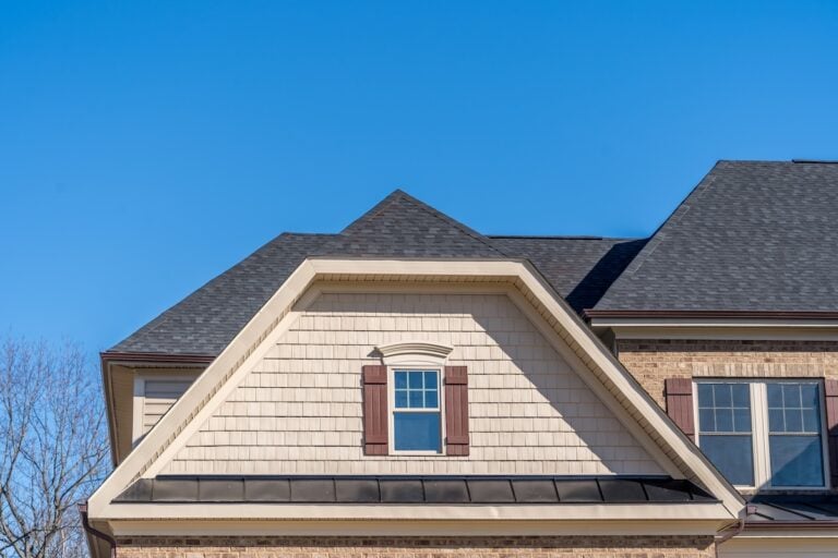 A close-up of a house with a dutch gable roof, dark shingles, beige siding, a single window with brown shutters, and clear blue sky in the background. Part of a leafless tree is visible on the left.