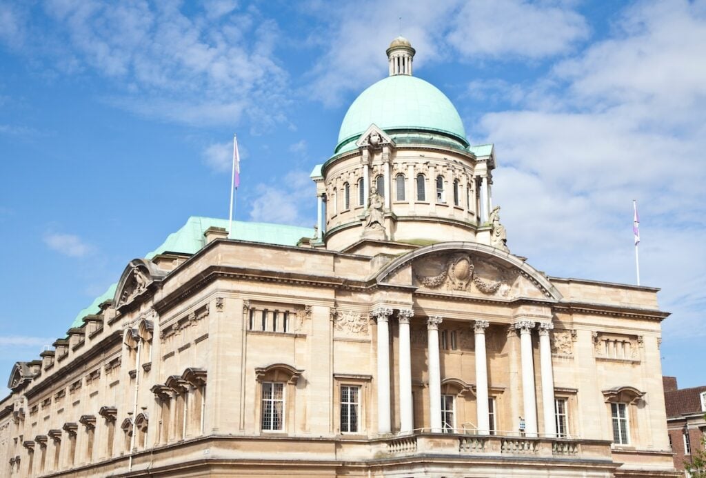 A historic building with a green dome roof, large columns, and ornate architectural details stands against a blue sky with scattered clouds. Two flagpoles frame the entrance, enhancing its grand appearance.