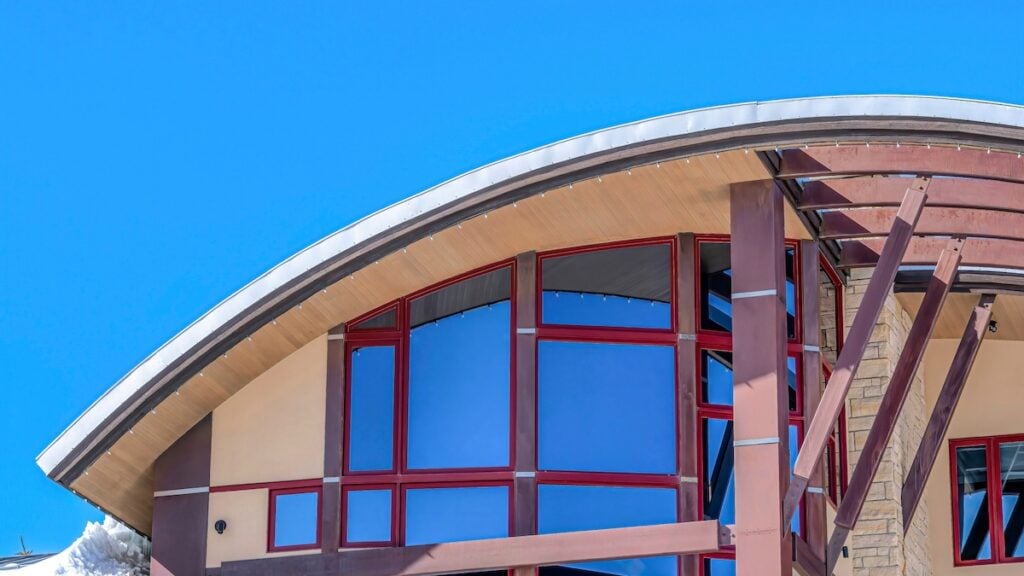 Modern building with a striking curved roof, large blue-tinted windows framed in red, and exposed wooden beams, set against a clear blue sky.