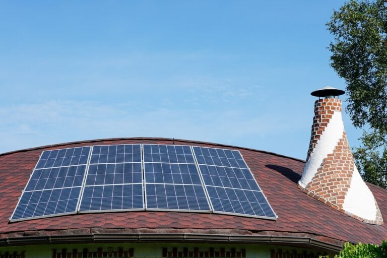 Solar panels are mounted on a curved roof with red shingles, set beside a unique brick chimney against a clear blue sky, while green foliage lines the roof’s edge.