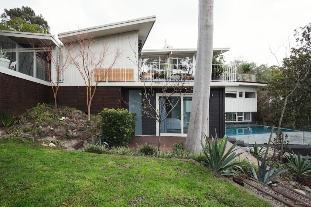 Modern two-story house with a striking butterfly roof, large windows, a balcony, and a swimming pool, surrounded by trees, shrubs, and a well-kept lawn, with a sloped garden and stone steps in the foreground.