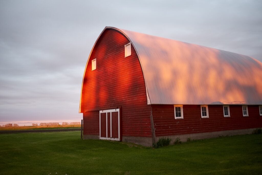 A red barn with a curved roof and white trim is illuminated by the warm light of a sunset, standing on green grass with a cloudy sky in the background.