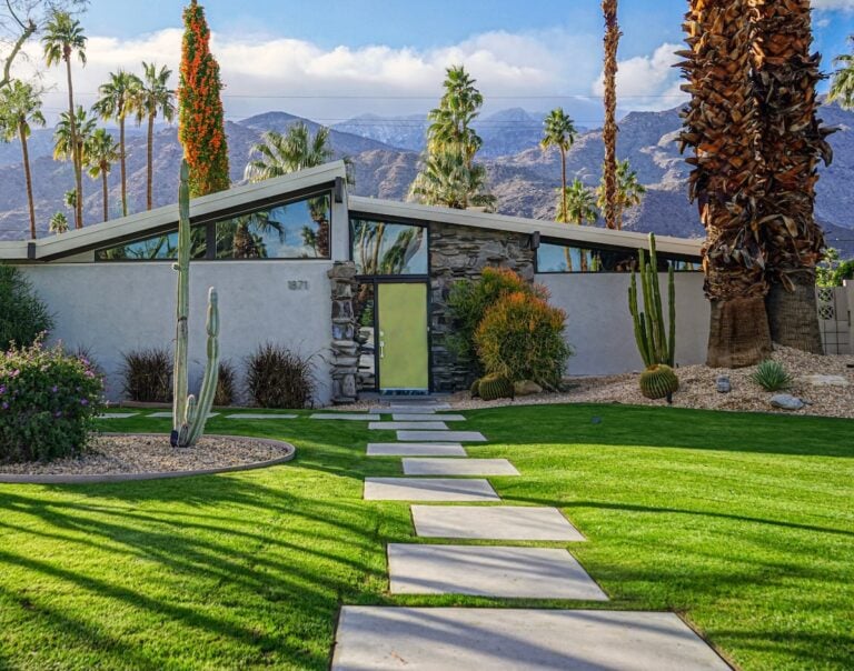 A modern, mid-century house with a striking butterfly roof, large glass door, and stone details. Lush green lawn, palm trees, and cacti fill the yard; mountains and blue sky create a stunning backdrop.