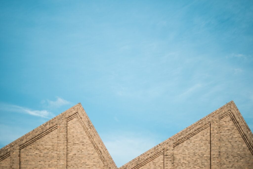 Two triangular brick rooftops, reminiscent of a sawtooth roof, stand against a clear blue sky with a few wispy clouds. The image highlights the geometric shapes and clean architectural lines.
