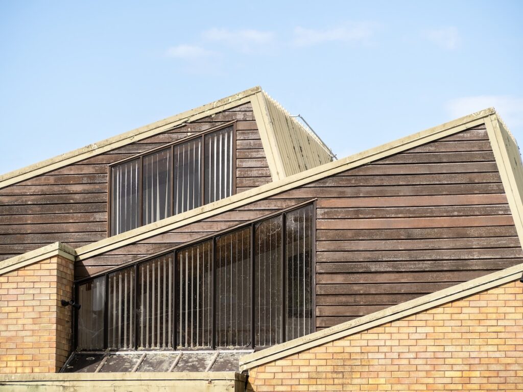 Angular modern building with wooden and brick exterior, featuring large vertical windows and overlapping slanted saltbox roofs against a clear blue sky.