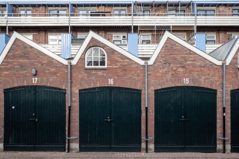 Three adjoining brick garages with green doors numbered 17, 16, and 15 sit below a residential building with balconies, metal railings, and a saltbox roof. The middle garage features a small arched window for added charm.