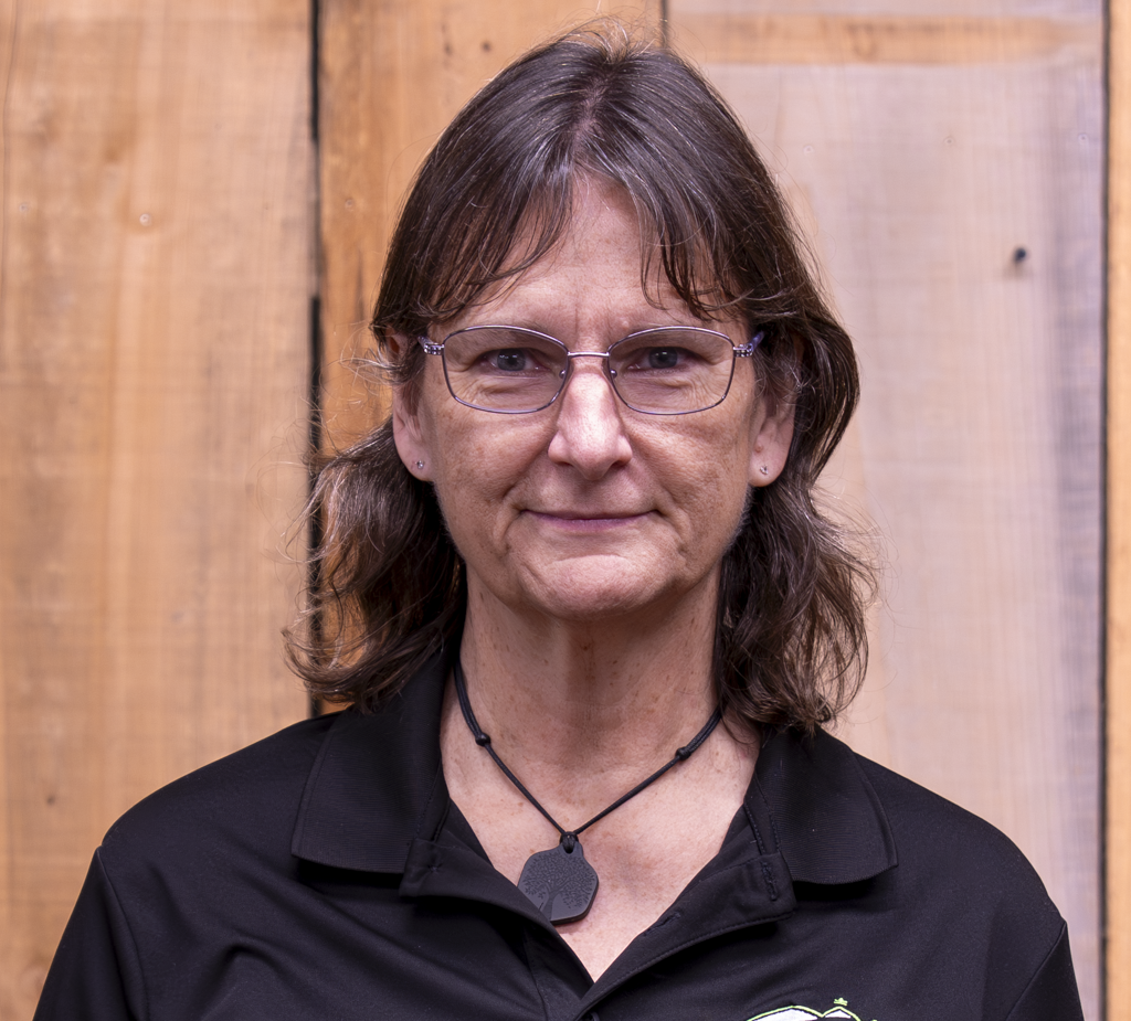 A woman with shoulder-length brown hair and glasses stands in front of a wooden background, wearing a black collared shirt and a black necklace with a rectangular pendant.