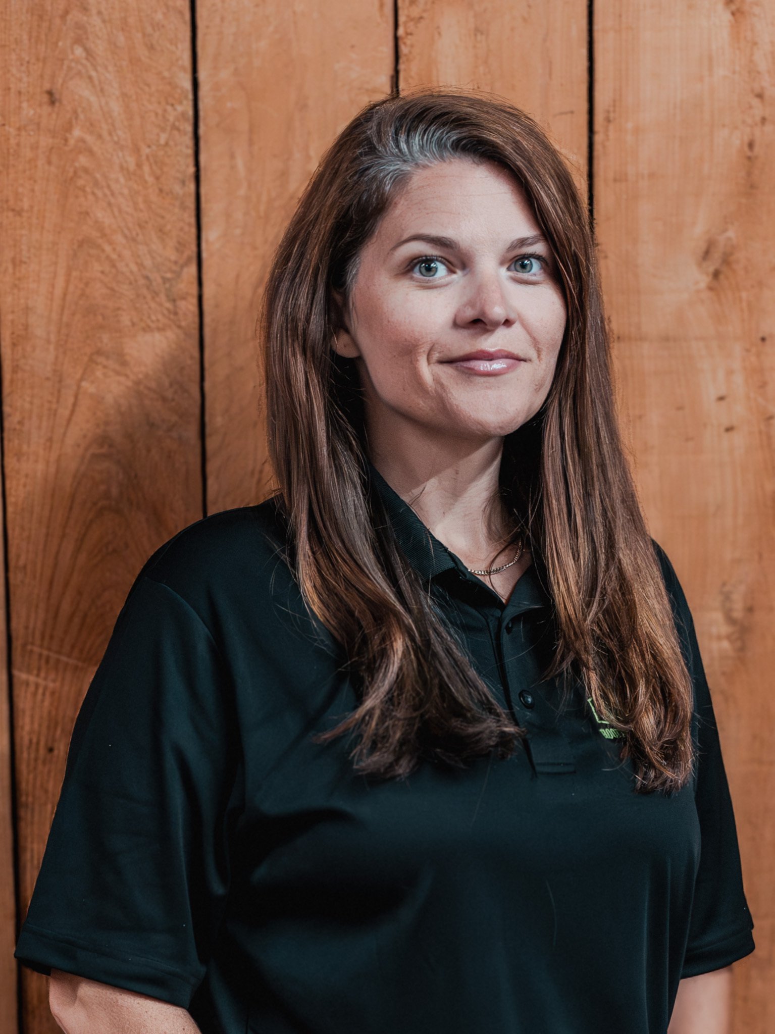 A woman with long brown hair wearing a black collared shirt stands in front of a wooden wall, looking at the camera with a slight smile.