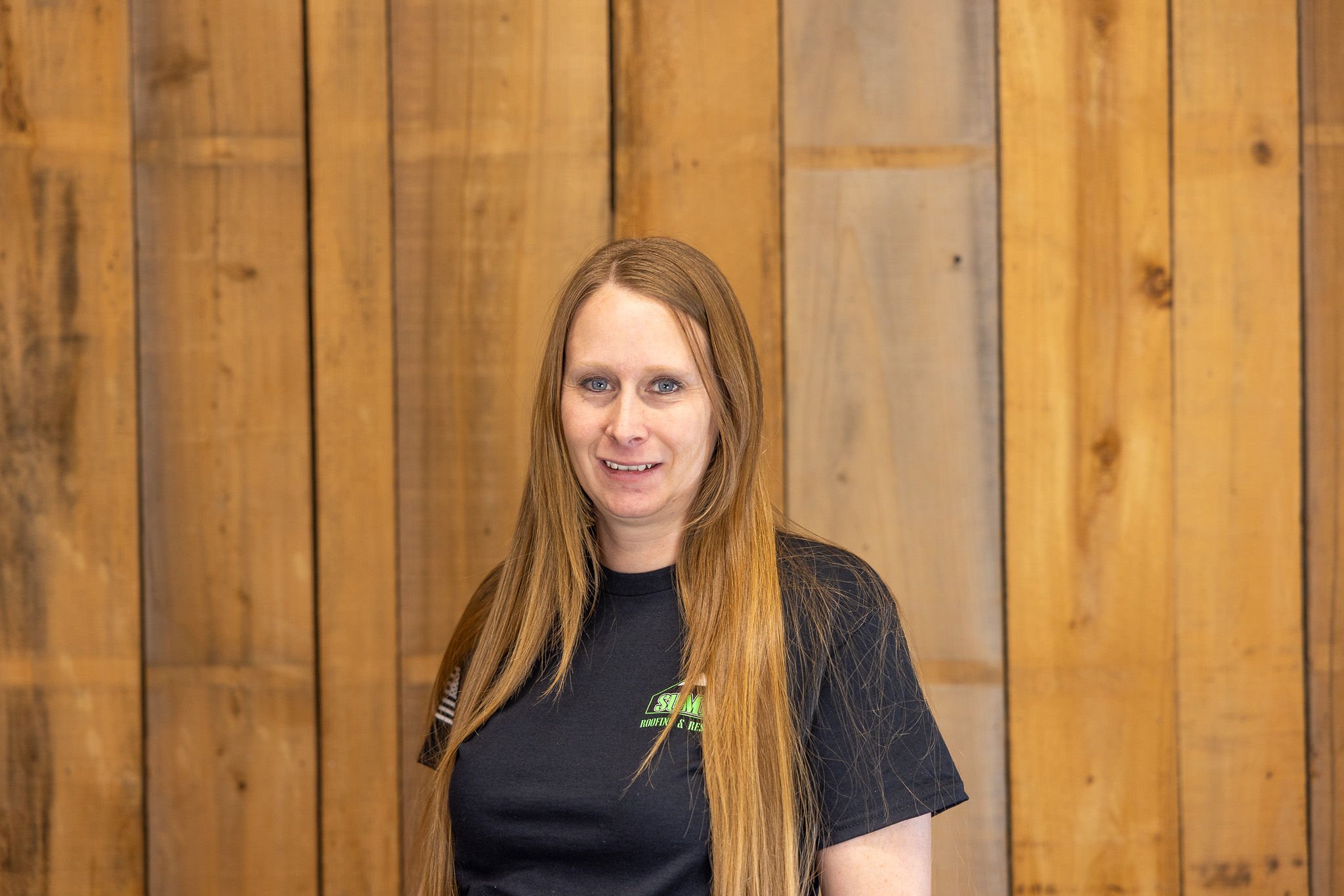 A woman with long straight blonde hair wearing a black t-shirt stands in front of a wooden plank wall, smiling slightly at the camera.