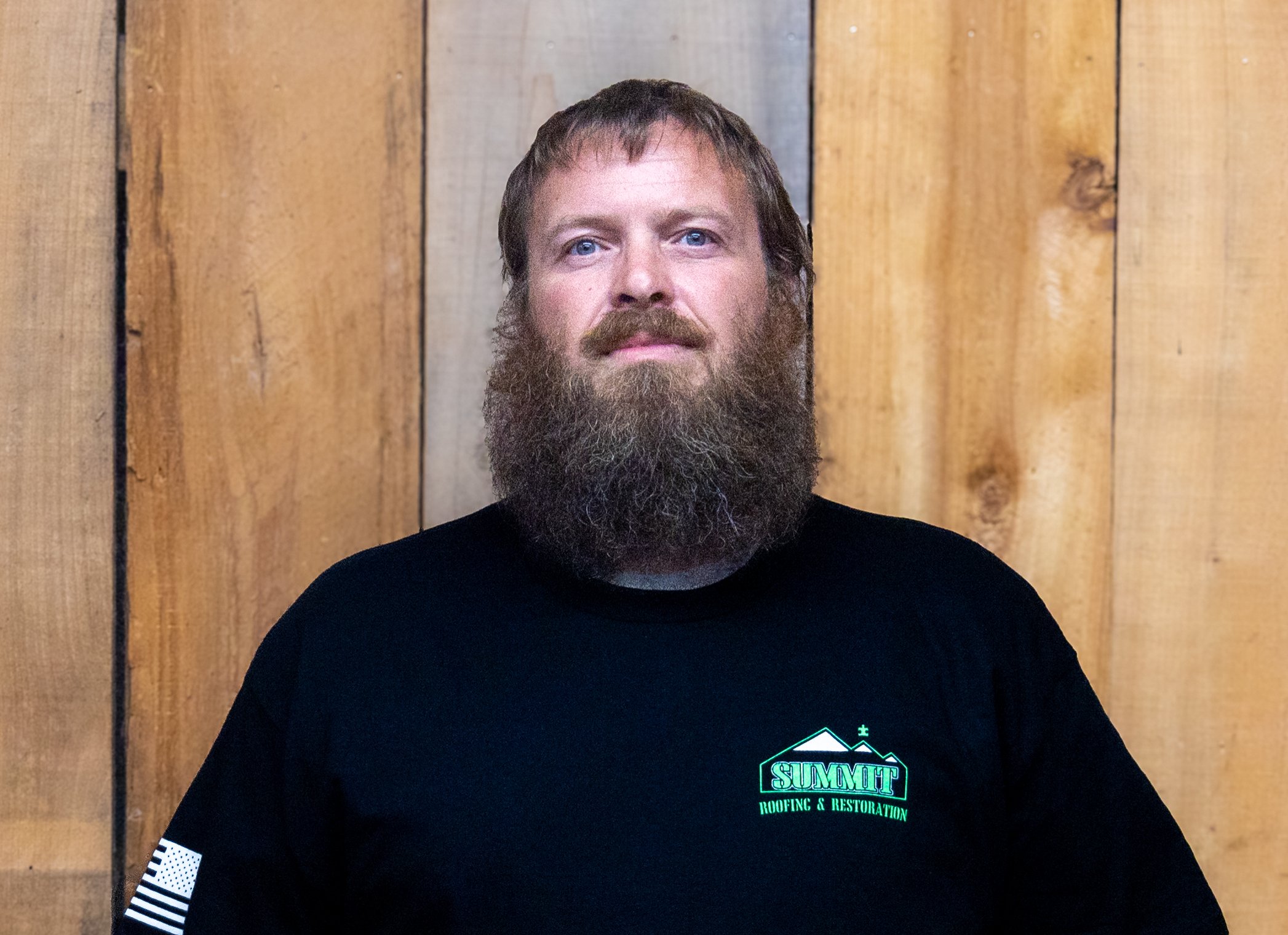 A man with a full brown beard and mustache stands in front of a wooden plank wall, wearing a black 