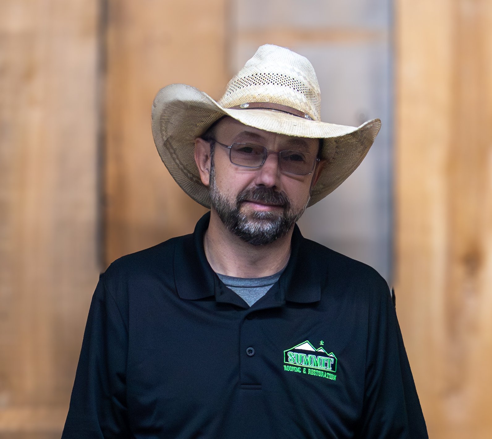 A man with a beard and glasses wears a straw cowboy hat and a black polo shirt with a green 