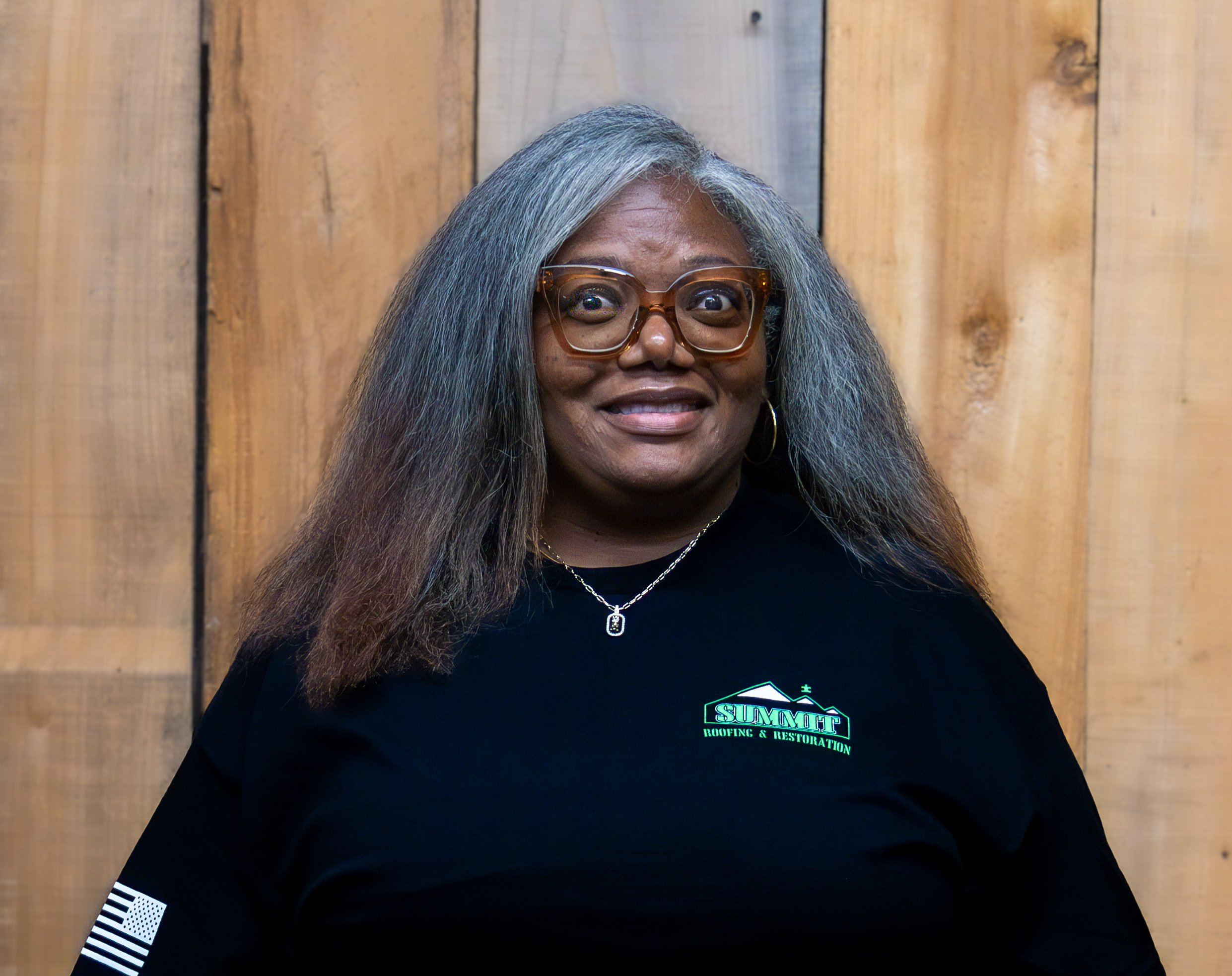 A woman with long gray hair and large glasses smiles at the camera. She wears a black t-shirt with a Summit logo and stands in front of a wooden background.