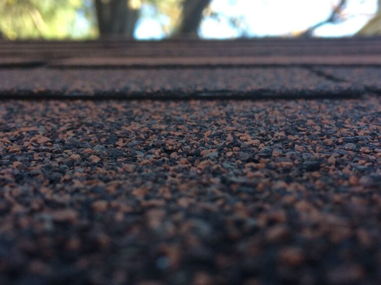 Close-up of brown and black asphalt roof shingles with a blurred background of tree branches and sky.