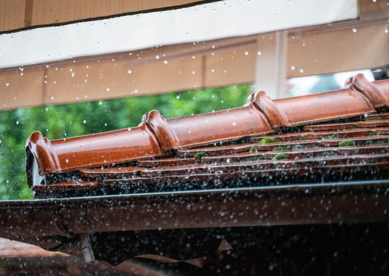 Rain falls on a sloped, reddish-brown tiled roof with moss growing between the tiles. Water droplets splash and blur in the foreground, while green foliage is visible in the background.