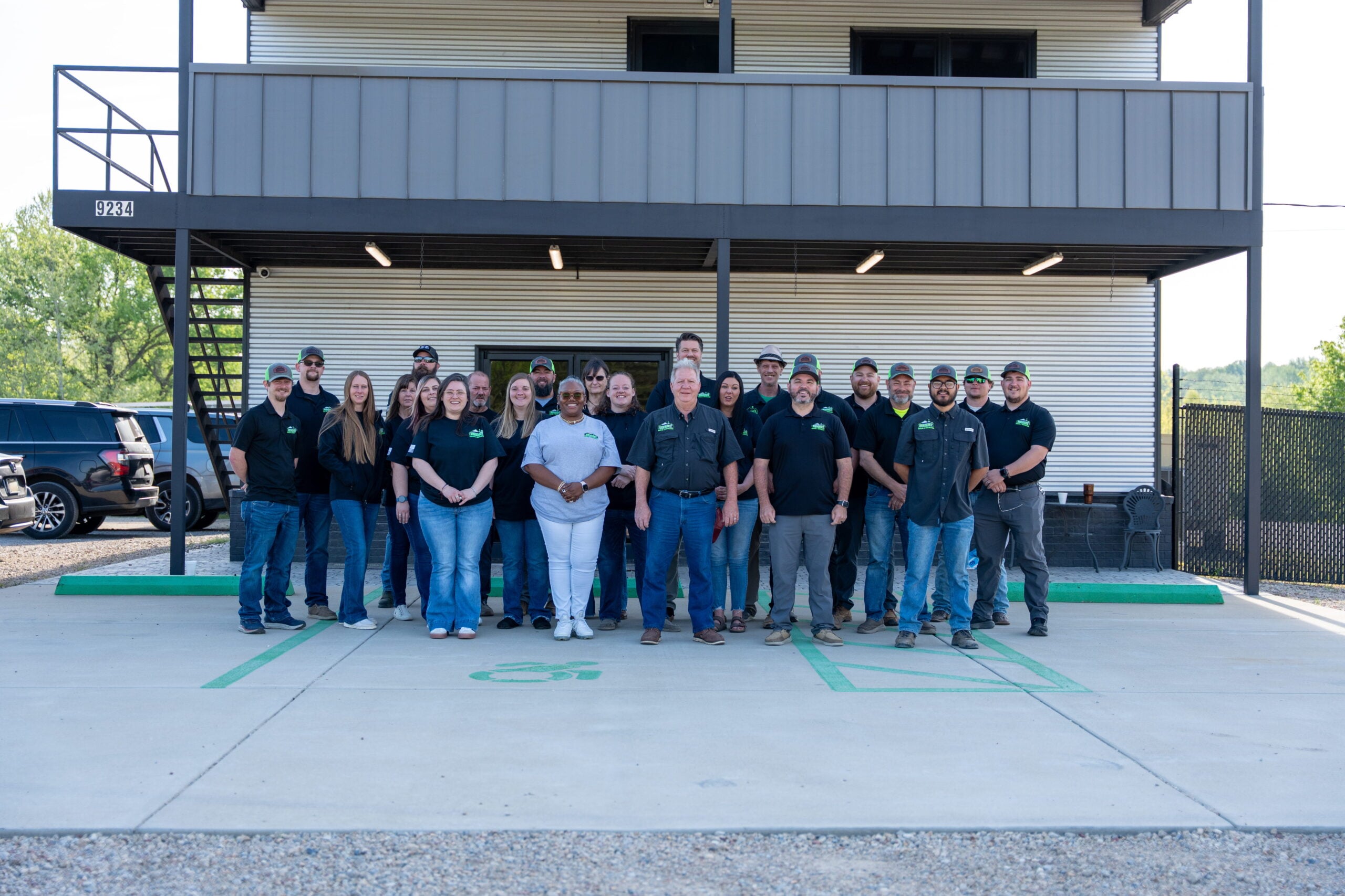 A group of 22 people pose together in front of a two-story building, standing on a concrete lot with a green handicap parking symbol visible in front.