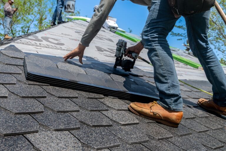 A person in jeans and brown shoes installs asphalt shingles on a roof using a nail gun, with a partially completed roof and blue sky in the background.