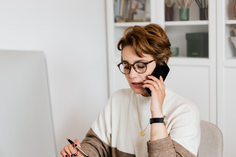 Woman with short hair and glasses talking on a smartphone while holding a pen, sitting at a desk with a computer in a home office setting. Shelves with books and decor are in the background.